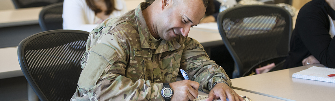 student in military attire smiling and completing paperwork at a desk