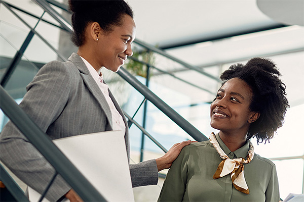 two smiling people in professional attire standing on stairs. one person's hand rests on the other's shoulder.