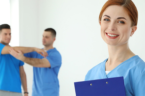 physical therapy aide holding clipboard stands in front of patient receiving physical therapy