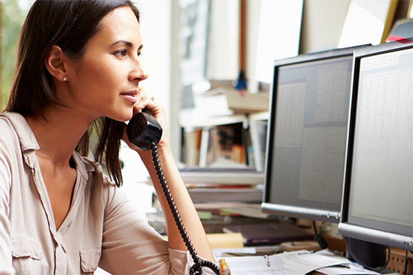person listening to phone in front of spreadsheets on computer monitors