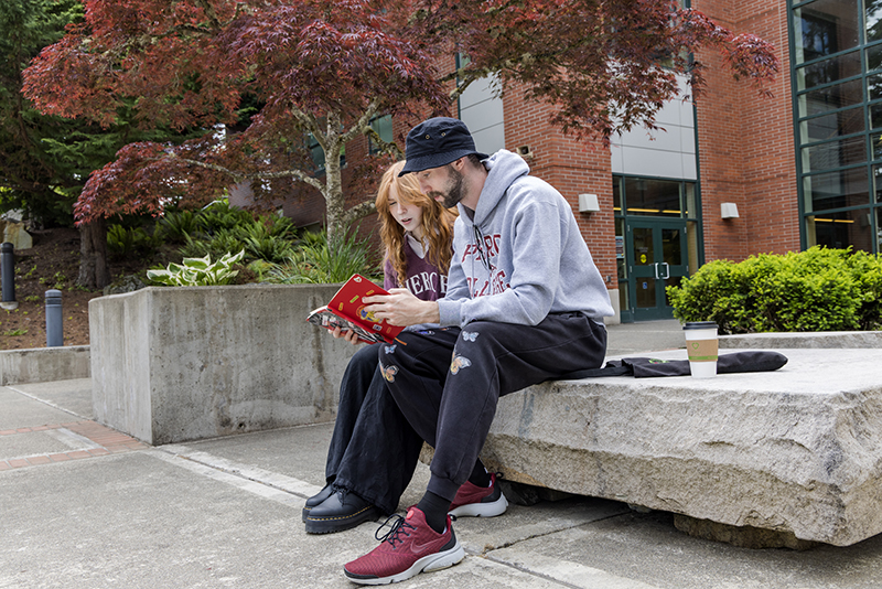 two students read a book together outside of the college center building