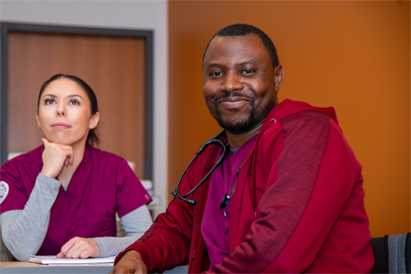 two nursing students wearing scrubs in classroom watching presentation