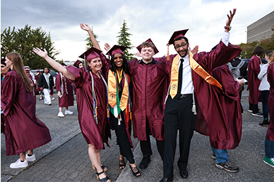 Four college graduates in maroon caps and gowns pose joyfully with arms raised, smiling widely. Two wear honor stoles, and the group stands outdoors among other graduates under a cloudy sky.
