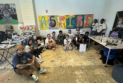 A diverse group of individuals sit and kneel together in an art studio, smiling at the camera. Behind them is a colorful mural spelling out 
