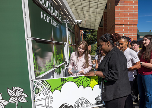 Group of diverse people gather around tall, green food lockers decorated with the GoodRoots Northwest Logo
