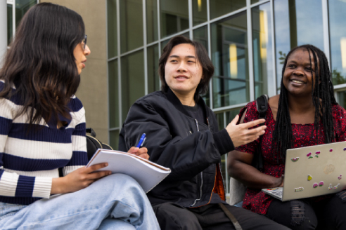 Three students engage in a discussion outdoors, with one taking notes and another using a laptop, surrounded by modern architecture.