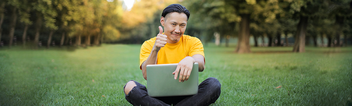 Man with laptop sitting on grass in clearing, making thumbs up sign