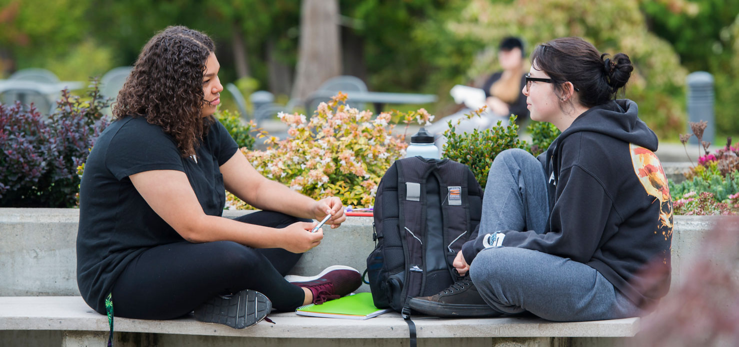 two students sitting on a concrete bench on campus having a conversation