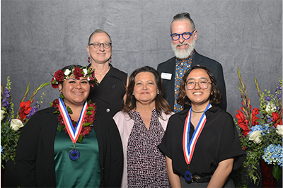 Two award recipients wearing medals stand, smiling proudly. One wears a green dress and a cultural headdress; the other wears glasses and a black outfit. Behind them, three administrators pose supportively. The group stands together against a neutral gray background adorned with vibrant red, blue, and white floral arrangements.