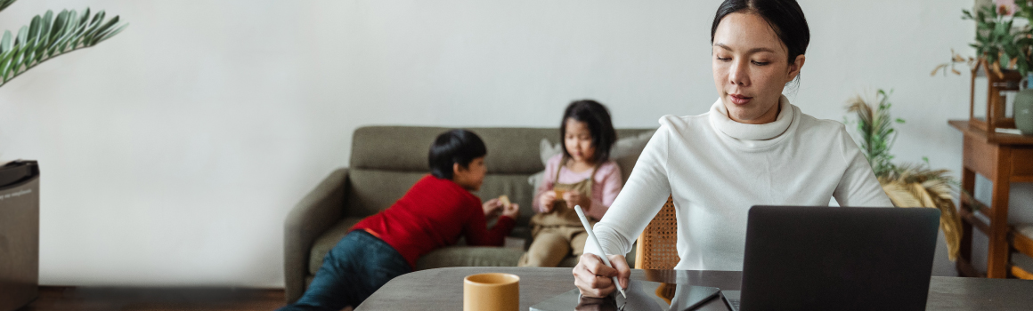 Woman using laptop at table while children play on couch in background