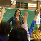 teacher standing in front of classroom where children are raising their hands