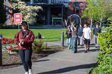 Woman with dark hair and red Pierce College jacket holds a sign that says Tour Guide as she leads visitors through campus